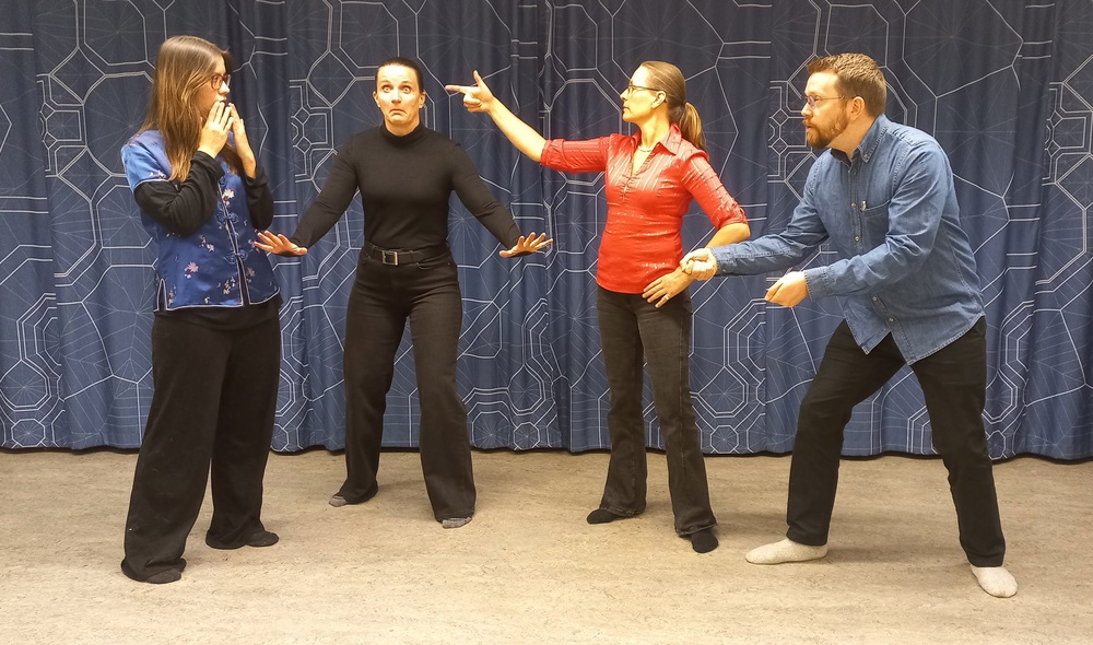 Fyra personer står i en grupp framför ett blått draperi och gestikulerar mot varandra, som i en teaterövning.