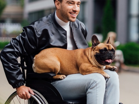 En leende man i rullstol utomhus med en fransk bulldogg i knät.