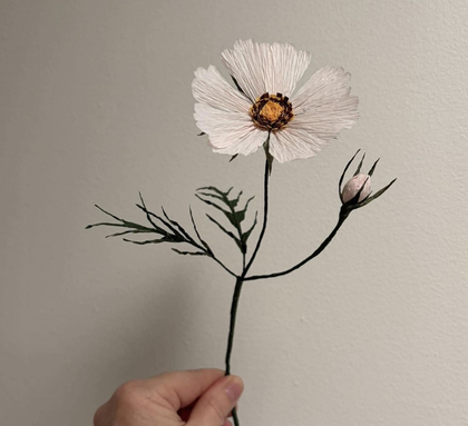 En hand håller en konstgjord blomma med ljusa kronblad och en knopp mot en ljus bakgrund.