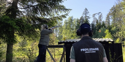 Två personer skjuter lerduvor i en skogsmiljö, en med gevär och en med väst märkt 'Provledare'.