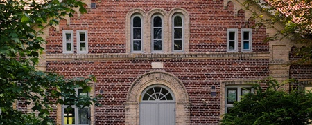 En gammal tegelbyggnad med spetsigt tak, sedd från en träbro omgiven av grönska.