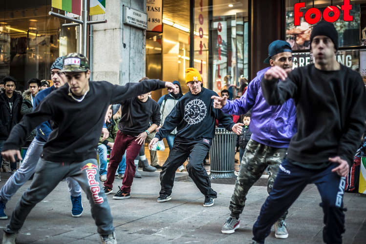En grupp unga män dansar hiphop på en stadsgata, omgivna av åskådare och butiker.