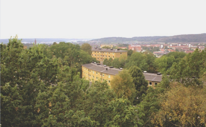 Vy över bostadshus omgivna av grönskande träd med stad och kullar i bakgrunden.