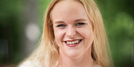 Leende ung kvinna med långt blont hår och vit blus står utomhus med suddig grön bakgrund.