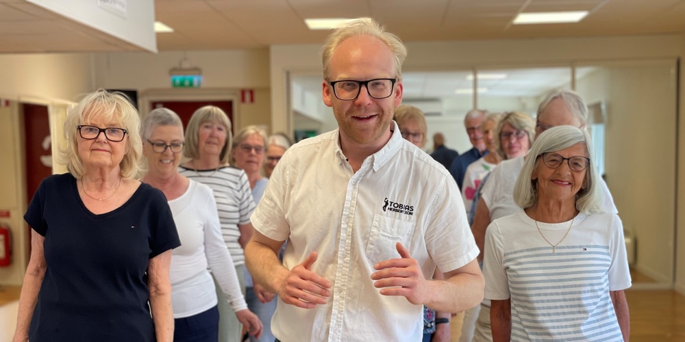 En grupp äldre personer och en yngre man står tillsammans inomhus, leende och avslappnade.