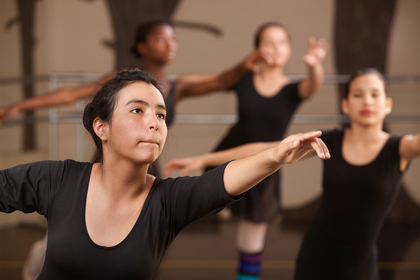 Fyra unga personer i svarta danskläder övar balett i en dansstudio, fokus på en person i förgrunden.