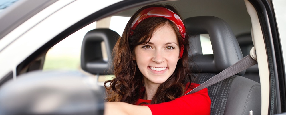 Ung kvinna med långt brunt hår och röd tröja sitter leende bakom ratten i en bil.