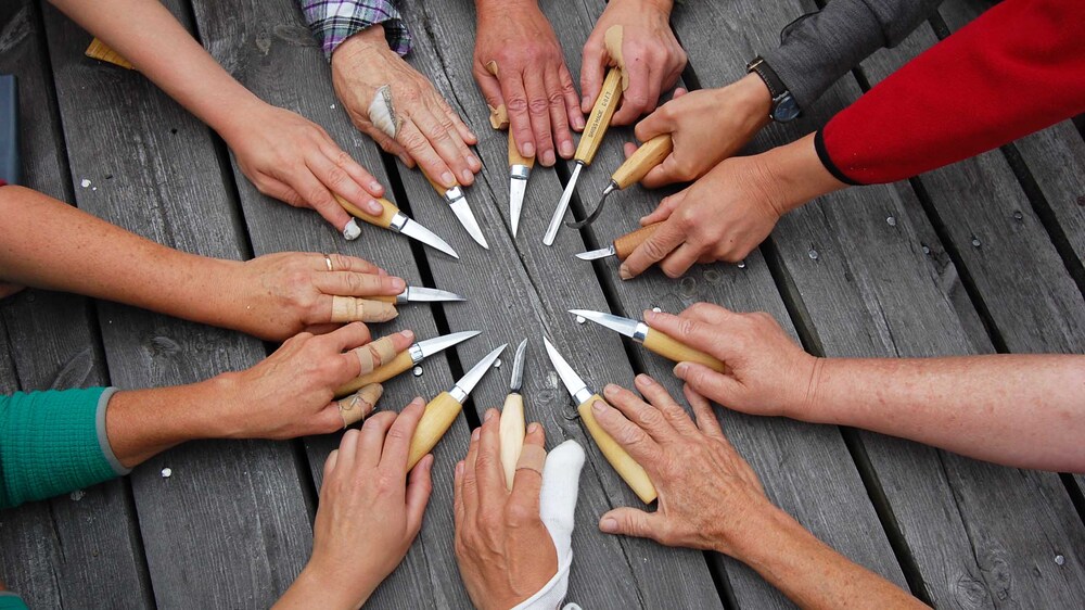 Flera händer håller knivar i en cirkel på ett träbord utomhus.