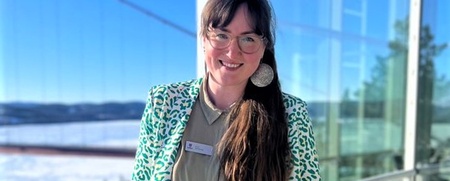 Kvinna med långt hår och glasögon står leende på en solig balkong med vinterlandskap i bakgrunden.