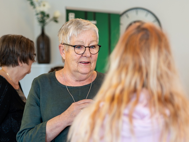 Äldre kvinna med glasögon samtalar med en yngre person i ett ljust rum med växter och klocka på väggen.