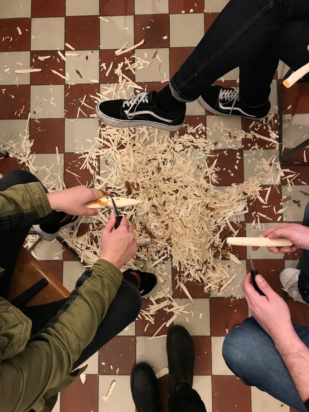 Tre personer täljer träpinnar och träspån ligger utspridda på ett rutigt golv.