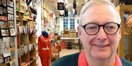 Äldre man med glasögon ler i ett rum fullt av samlarföremål och memorabilia.