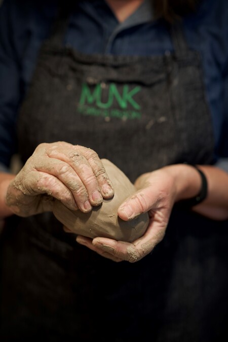 En person formar lera med händerna, iförd förkläde med texten 'MUNK'.
