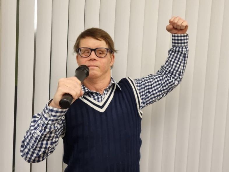 Person med glasögon håller en mikrofon och höjer ena armen framför vita persienner.