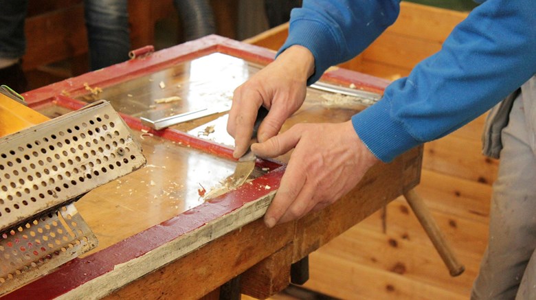 En person skrapar vax från en bikupa-ram med ett verktyg på ett träbord.