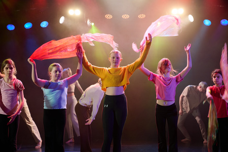 En grupp unga personer dansar på scen med färgglada sjalar under scenbelysning.