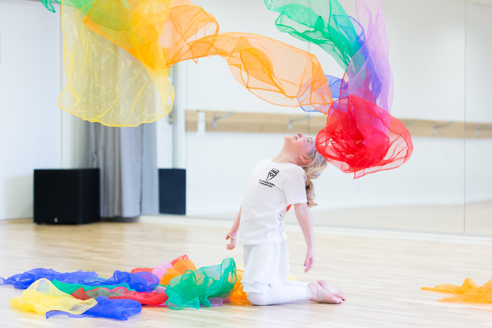 Barn i vit klädsel knäar på golvet i en dansstudio och kastar färgglada tygsjok i luften.