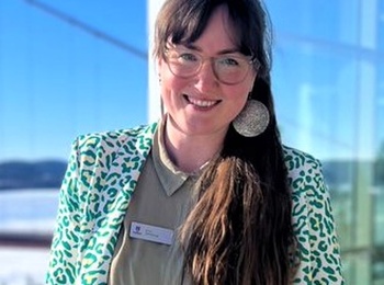 Kvinna med långt hår och glasögon står leende på en solig balkong med vinterlandskap i bakgrunden.