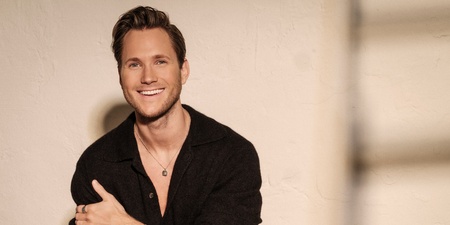 Man sitter mot en ljus vägg, klädd i svart skjorta och byxor, med armarna korsade över bröstet.