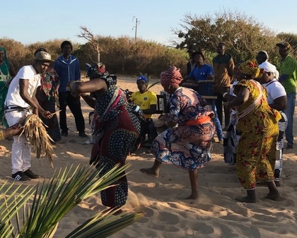 En grupp människor dansar och spelar musik på en sandig plats utomhus, klädda i färgglada traditionella kläder.