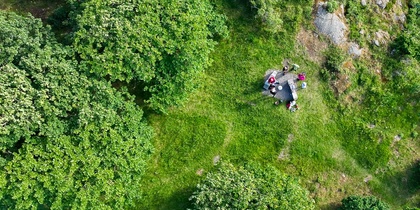 Flygbild över en grönskande glänta med några personer som sitter på en filt mitt i naturen.