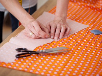 Händer som nålar fast mönsterpapper på orange tyg med sax och krita bredvid.