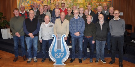 Gruppfoto av män i flera rader framför en färgglad väggbonad, med en blå symbol i förgrunden.
