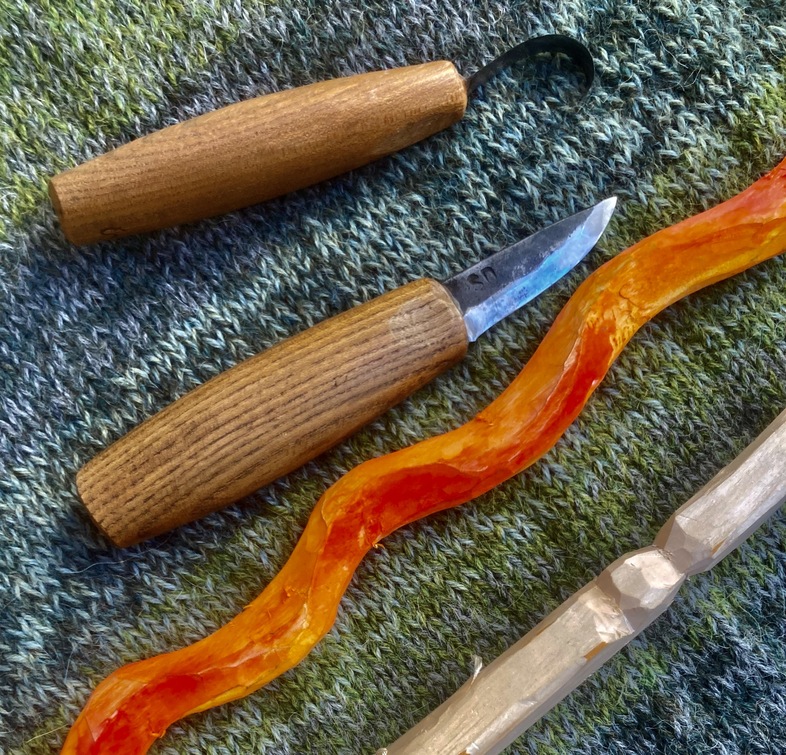 Två träsnidarverktyg och två snidade träpinnar ligger på en grön yta.