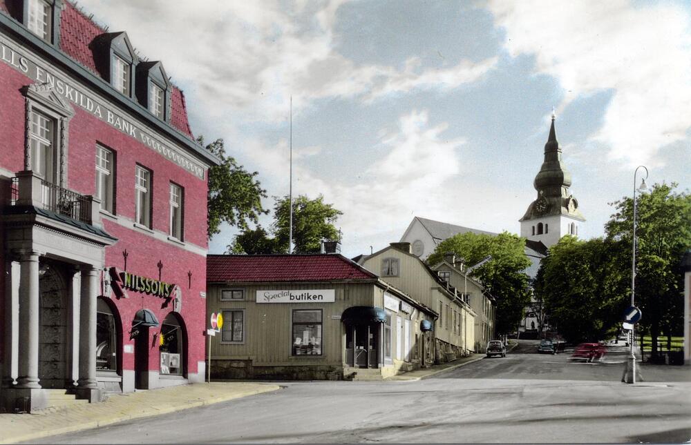Stadsgata i Hudiksvall med äldre byggnader, bank, butik och kyrka i bakgrunden, några bilar på vägen.