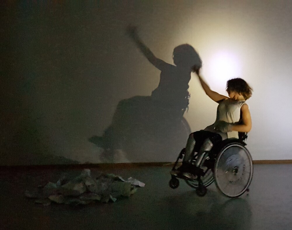 En person i rullstol sträcker upp armen mot en vägg där deras skugga syns tydligt. En hög med papper ligger på golvet.