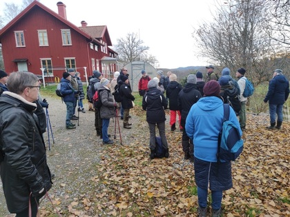 En grupp människor står utomhus vid ett rött trähus, klädda för höstväder, och lyssnar på en guide.