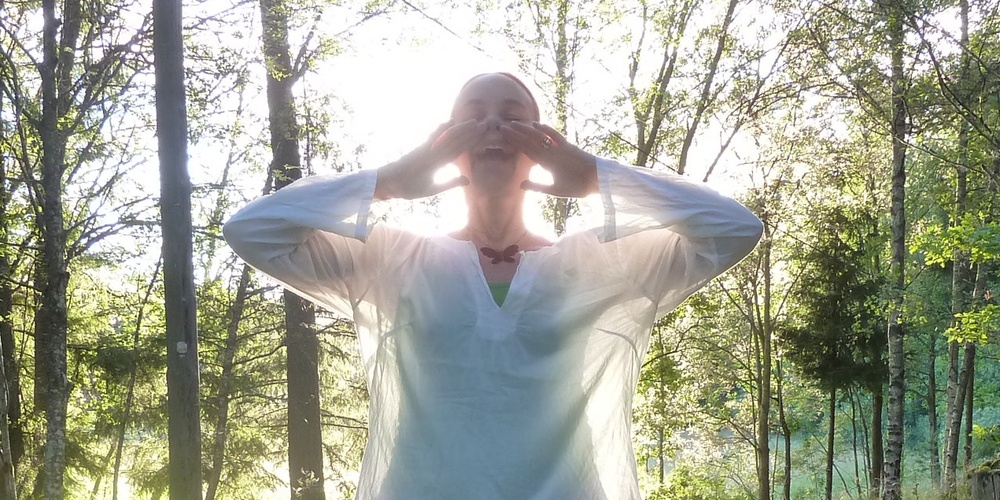 Person i vit tunika står i en solig skog med händerna vid huvudet.