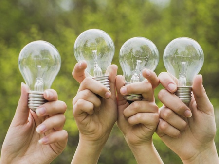 Fyra händer håller upp varsin glödlampa utomhus med grönska i bakgrunden.