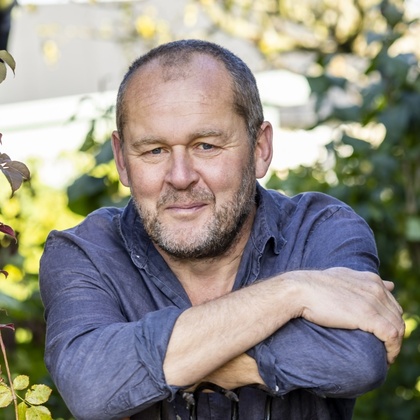 En person står i en trädgård med armarna vilande på ett trädgårdsredskap, omgiven av gröna växter.