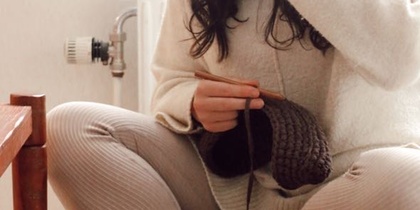 Person sitter korslagd på en kudde och stickar med mörkbrunt garn i ett ljust, mysigt rum.