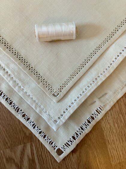 Tre beige linneservetter med dekorativa hålsömmar och en vit trådrulle ovanpå, liggande på ett trägolv.