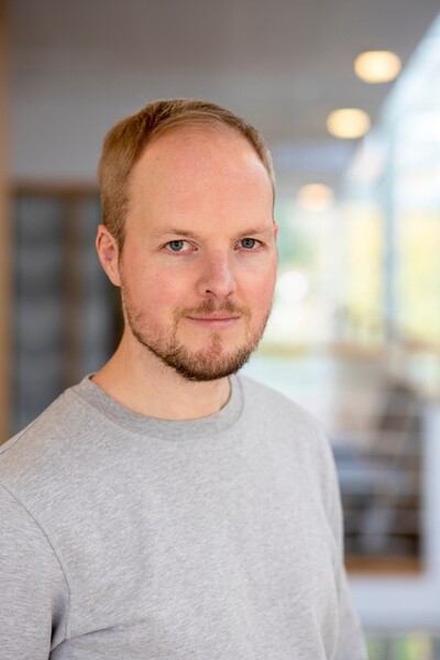 Man med kort hår och skägg bär en grå tröja och står inomhus i ett ljust, modernt rum.