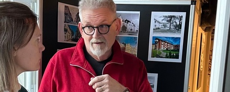 Två personer samtalar framför en utställningstavla med arkitekturbilder i en inomhusmiljö.