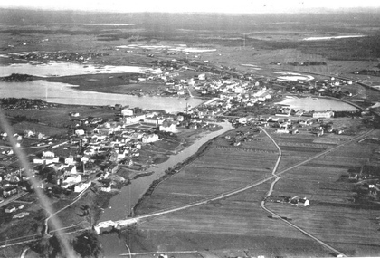 Svartvit flygbild över en liten stad med flod, broar, hus och omgivande åkrar.