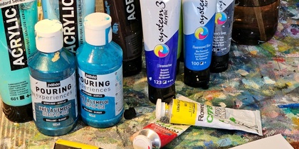 Ett bord med akrylfärger, penslar, markers, en sprayflaska och en teckning av en fågel framför en målning av påfåglar.