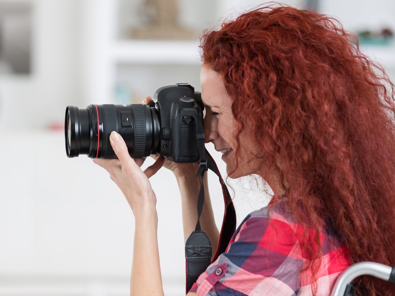 En person med långt rött hår sitter i rullstol och fotograferar med en systemkamera.