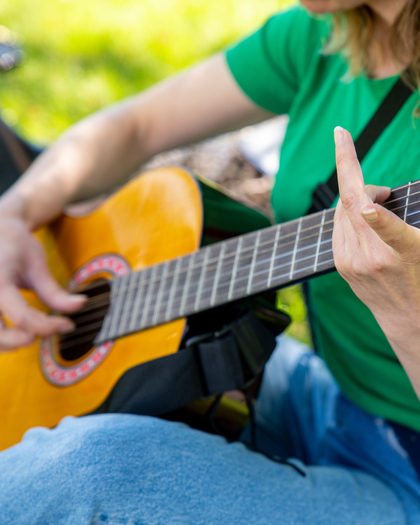 Person i grön tröja spelar akustisk gitarr utomhus, fokus på händer och gitarr.