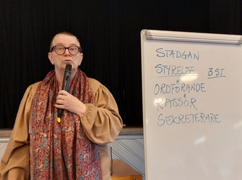 Person står vid ett blädderblock i en sal och håller en mikrofon. Text på blocket handlar om styrelseposter.