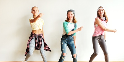 Tre ungdomar dansar energiskt i ett ljust rum med trägolv och vit vägg.