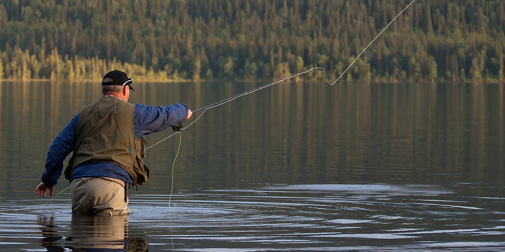En person står i vattnet och flugfiskar vid en sjö med skogsklädda berg i bakgrunden.