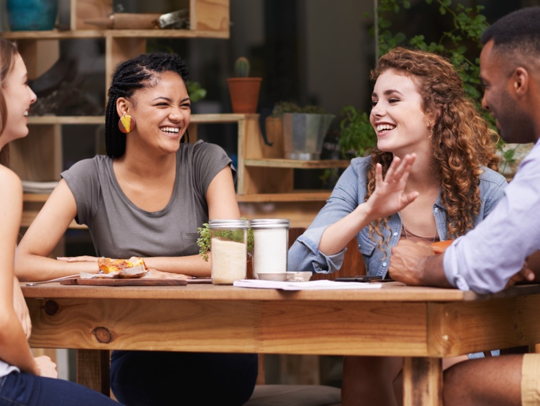 Fyra personer sitter vid ett träbord utomhus och samtalar, med växter i bakgrunden.