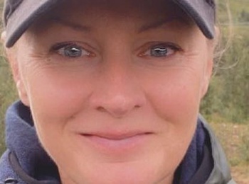 Kvinna i grön jacka och svart keps tar en selfie utomhus i naturmiljö.