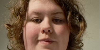 Person med lockigt, blont hår och vit t-shirt sitter framför en neutral bakgrund. Ansiktet är suddat.