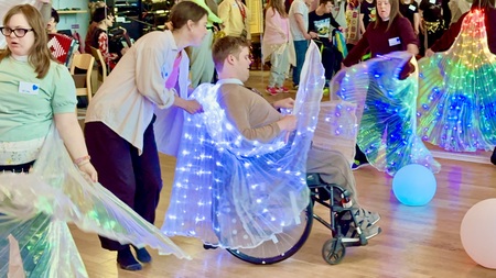 # personer i ett rum varav en sitter i en rullstol. De dansar med färggladda isisvingar. En fjärde person hjälper personen i rullstolen