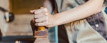 En person i arbetskläder hyvlar trä i en snickarverkstad, fokus på händer och verktyg.